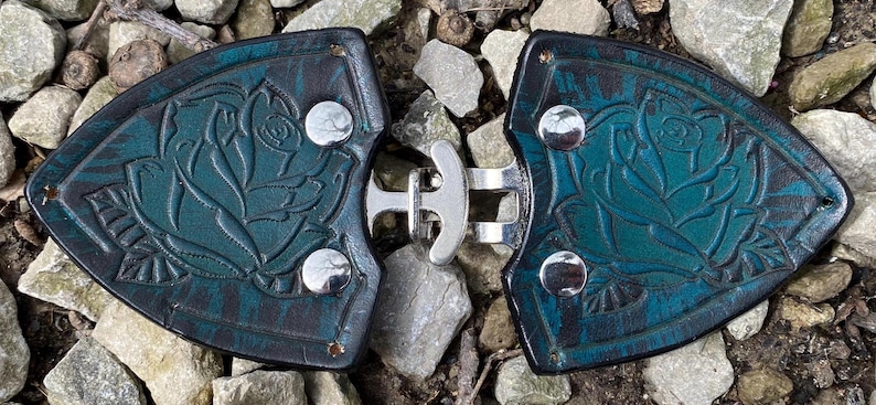Pu&ograve; includere: Un paio di fibbie per cintura in pelle blu turchese con un design a rosa e chiusure in metallo argentato.