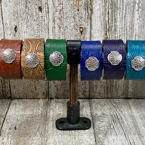May include: Ten leather bracelets with a Celtic knot design and a silver metal Celtic cross charm. The bracelets are displayed on a wooden stand and come in a variety of colors, including black, red, brown, tan, green, blue, and purple.