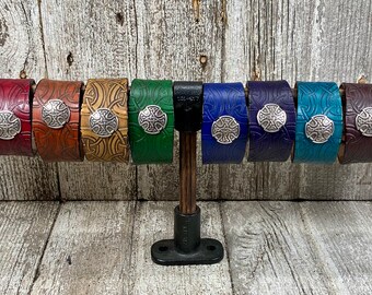May include: Ten leather bracelets with a Celtic knot design and a silver metal Celtic cross charm. The bracelets are displayed on a wooden stand and come in a variety of colors, including black, red, brown, tan, green, blue, and purple.