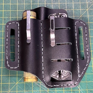 May include: Black leather multi-tool sheath with white stitching. The sheath has two compartments, one holding a brass-colored object and the other a multi-tool. Metal clips secure the items. The sheath is designed for carrying tools.