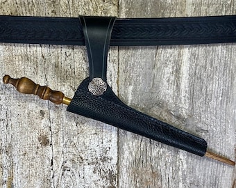 May include: A black leather sheath with a cross-shaped metal accent, attached to a black leather belt. A wooden stick is partially inserted into the sheath.