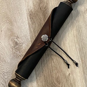 May include: A brown leather scroll case with a black fabric lining and a silver Celtic knot design on the front. The case has two black cords with silver beads at the ends.