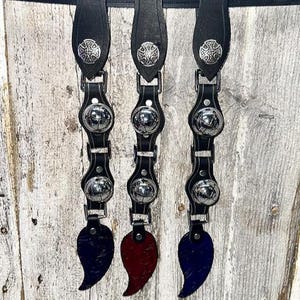 May include: Three black leather straps with silver-colored metal accents and decorative cross emblems. Each strap features three shiny silver spheres and a teardrop-shaped accent in either navy, red, or blue. The straps are hanging against a weathered wood background.