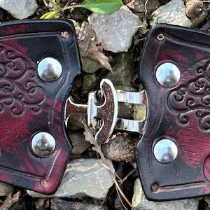 Pu&ograve; includere: Un paio di fibbie per cintura in pelle con un design ad albero della vita. Le fibbie sono di colore bordeaux e nero con accenti argentati.