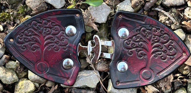 Pu&ograve; includere: Un paio di fibbie per cintura in pelle con un design ad albero della vita. Le fibbie sono di colore bordeaux e nero con accenti argentati.