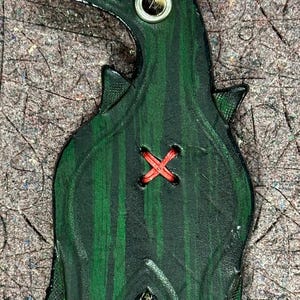 May include: A green leather dinosaur ornament with a red stitched X on the back. The ornament has a hole at the top for hanging with twine.