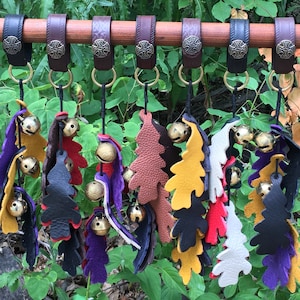 May include: A set of seven leather leaf-shaped wind chimes with bells hanging from a wooden dowel. Each chime is a different color: brown, black, purple, red, white, yellow, and orange.
