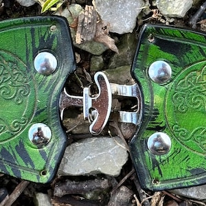 Pu&ograve; includere: Un paio di fibbie per cintura in pelle verde con un design ad albero della vita celtico e dettagli in metallo argentato.