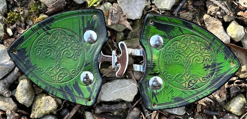 Pu&ograve; includere: Un paio di fibbie per cintura in pelle verde con un design ad albero della vita celtico e dettagli in metallo argentato.
