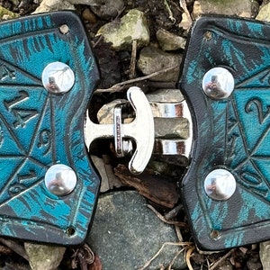 Pu&ograve; includere: Un paio di porta dadi in pelle nera con un design marmoreo blu turchese e nero. Ogni porta dadi presenta un dado a 20 facce con i numeri da 1 a 20 in rilievo. I porta dadi sono collegati da una fibbia in metallo argentato.