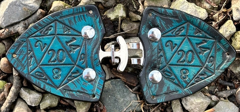 Pu&ograve; includere: Un paio di porta dadi in pelle nera con un design marmoreo blu turchese e nero. Ogni porta dadi presenta un dado a 20 facce con i numeri da 1 a 20 in rilievo. I porta dadi sono collegati da una fibbia in metallo argentato.