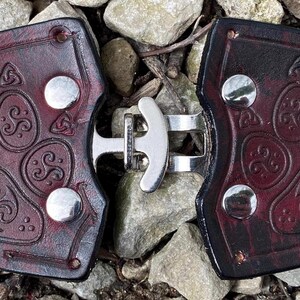Pu&ograve; includere: Un paio di fibbie per cintura in pelle marrone scuro con una chiusura in metallo argentato. Ogni fibbia presenta un design a forma di impronta di zampa in rilievo con nodi celtici.