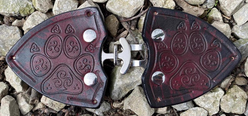 Pu&ograve; includere: Un paio di fibbie per cintura in pelle marrone scuro con una chiusura in metallo argentato. Ogni fibbia presenta un design a forma di impronta di zampa in rilievo con nodi celtici.