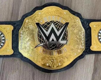 May include: A gold and black championship belt with the text "WORLD CHAMPION" and the WWE logo. The belt has a large center plate and two side plates, all with intricate designs and embellishments.