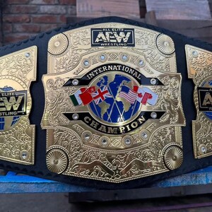 May include: A gold and black wrestling championship belt. The belt features the text "ALL ELITE AEW WRESTLING" on the side plates and "INTERNATIONAL CHAMPION" on the center plate. Flags of different countries are also displayed.