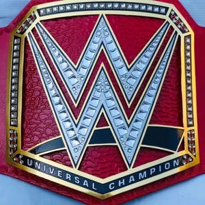 May include: A red and gold championship belt with the letters "W" in the center. The belt has a textured red background and the words "UNIVERSAL CHAMPION" are displayed on the bottom.