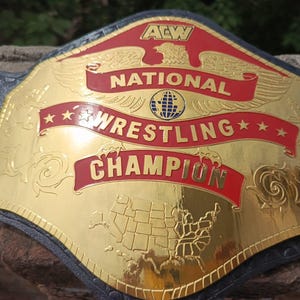May include: A gold and red wrestling championship belt with the text "National Wrestling Champion". The belt features a gold plate with a map of the United States and decorative scrollwork. The belt is black.