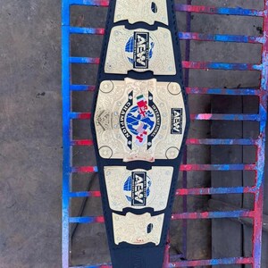 May include: A black wrestling championship belt with gold-colored plates and the text "AEW" and "INTERNATIONAL CHAMPION". The belt features a blue and white globe design and is displayed on a metal surface.