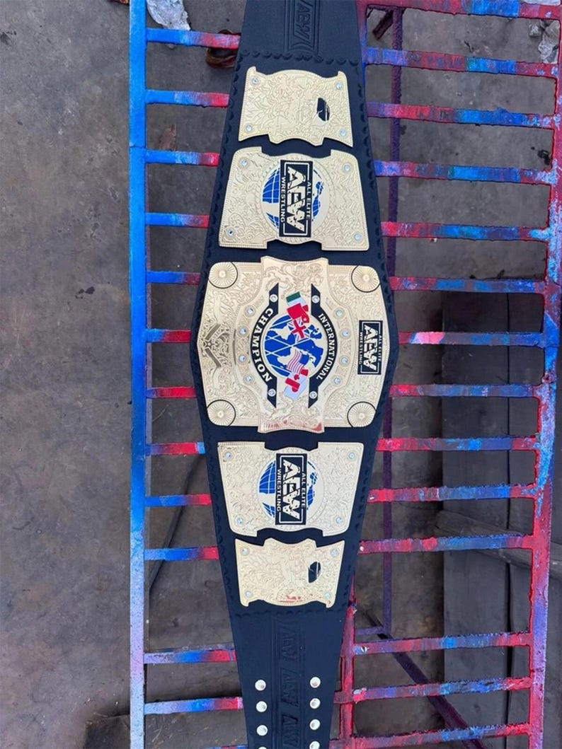 May include: A black wrestling championship belt with gold-colored plates and the text "AEW" and "INTERNATIONAL CHAMPION". The belt features a blue and white globe design and is displayed on a metal surface.