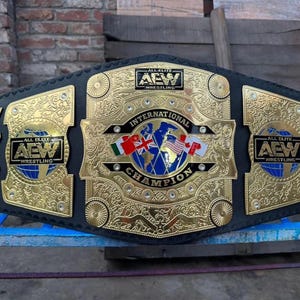 May include: A gold and black wrestling championship belt with the text "ALL ELITE WRESTLING" and "INTERNATIONAL CHAMPION". The belt features gold filigree and the flags of several countries.