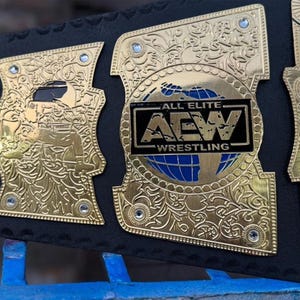 May include: Close-up of a gold and black wrestling championship belt. The belt features intricate gold detailing, including floral patterns and the text "ALL ELITE AEW WRESTLING" on a black and blue background. The belt is made of black leather.