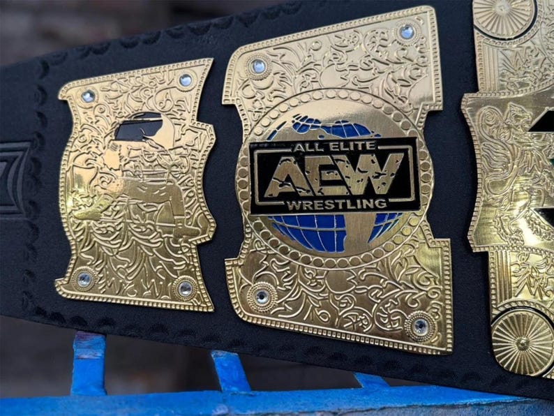 May include: Close-up of a gold and black wrestling championship belt. The belt features intricate gold detailing, including floral patterns and the text "ALL ELITE AEW WRESTLING" on a black and blue background. The belt is made of black leather.