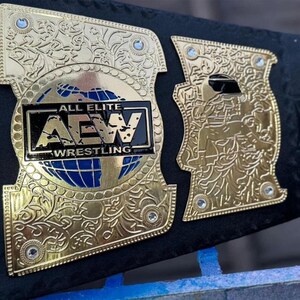 May include: Close-up of a gold and black wrestling championship belt. The belt features intricate gold detailing, including floral patterns and the text "ALL ELITE AEW WRESTLING" on the center plate. Blue accents are visible.