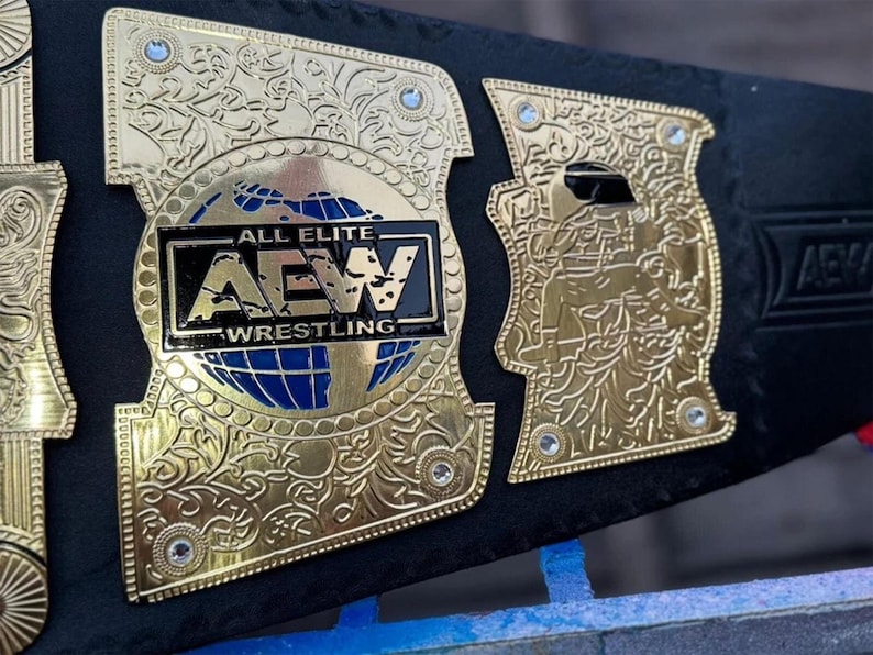May include: Close-up of a gold and black wrestling championship belt. The belt features intricate gold detailing, including floral patterns and the text "ALL ELITE AEW WRESTLING" on the center plate. Blue accents are visible.