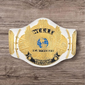 May include: A white and gold wrestling championship belt with the words "WORLD HEAVYWEIGHT WRESTLING CHAMPION" displayed. The belt features a gold eagle design with a blue globe in the center.