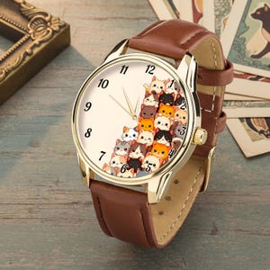 May include: A gold-toned wristwatch with a brown leather band. The watch face features a cream-colored background with black numerals and a playful design of stacked cartoon cats in various colors. The watch has gold hands.