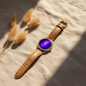 May include: A gold-toned wristwatch with a brown leather band. The watch face displays a vibrant purple and blue galaxy design. The watch is set against a neutral background with dried flowers.