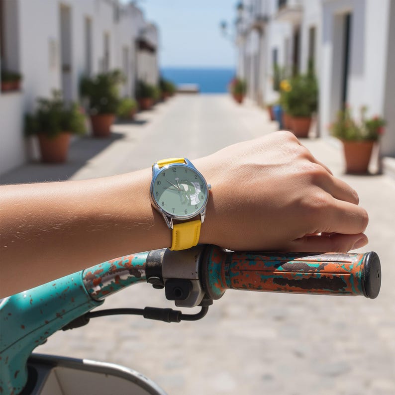 May include: A silver wristwatch with a light green face and a yellow leather band is worn on a wrist. The watch is held over a scooter handlebar. The background shows a street with white buildings and the ocean.