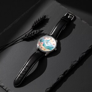 May include: A wristwatch with a black leather strap and a silver-tone circular case. The watch face features a colorful watercolor-style illustration of a mountain landscape with pink flowers, black numerals, and thin silver hands.