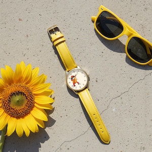 May include: A yellow wristwatch with a round face featuring a cartoon hot dog character dabbing, paired with a bright yellow sunflower and yellow-framed sunglasses. The watch has a smooth yellow leather strap and a gold-tone metal watch case.