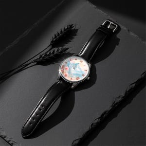 K&ouml;nnte beinhalten: Eine silberne Armbanduhr mit schwarzem Lederarmband. Das Zifferblatt zeigt ein Aquarelldesign in Rosa, Blau und Wei&szlig;. R&ouml;mische Ziffern markieren die Stunden. Die Uhr liegt auf einer dunklen Oberfl&auml;che mit einem Weizen&auml;hrenzweig.
