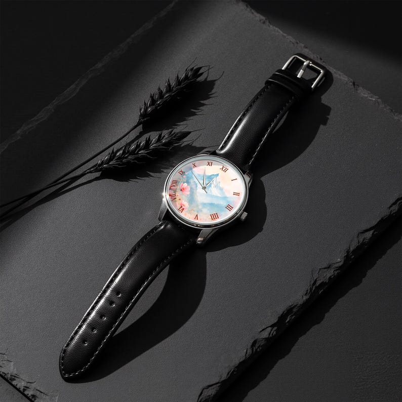 K&ouml;nnte beinhalten: Eine silberne Armbanduhr mit schwarzem Lederarmband. Das Zifferblatt zeigt ein Aquarelldesign in Rosa, Blau und Wei&szlig;. R&ouml;mische Ziffern markieren die Stunden. Die Uhr liegt auf einer dunklen Oberfl&auml;che mit einem Weizen&auml;hrenzweig.