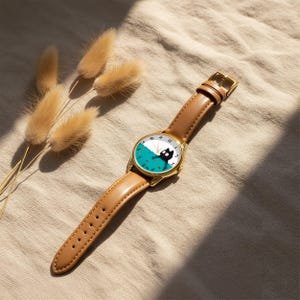 Pu&ograve; includere: Un orologio da polso color oro con cinturino in pelle marrone. Il quadrante presenta un design di gatto nero su sfondo turchese e bianco. L'orologio &egrave; appoggiato su un tessuto dai toni neutri con piante essiccate.