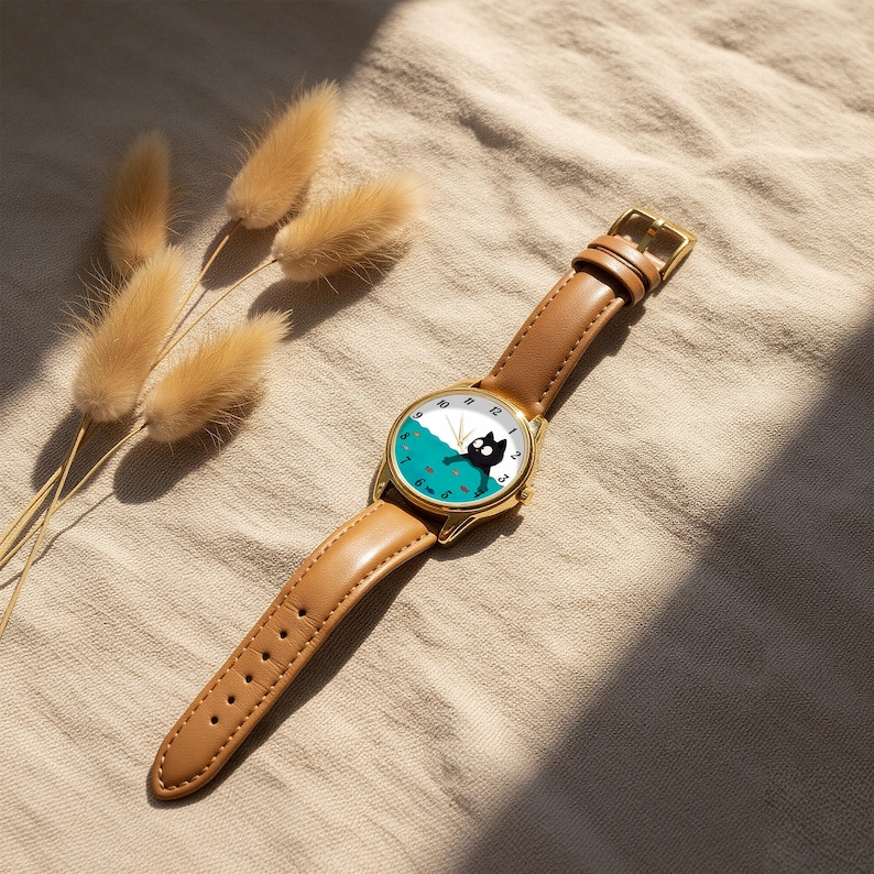 Pu&ograve; includere: Un orologio da polso color oro con cinturino in pelle marrone. Il quadrante presenta un design di gatto nero su sfondo turchese e bianco. L'orologio &egrave; appoggiato su un tessuto dai toni neutri con piante essiccate.