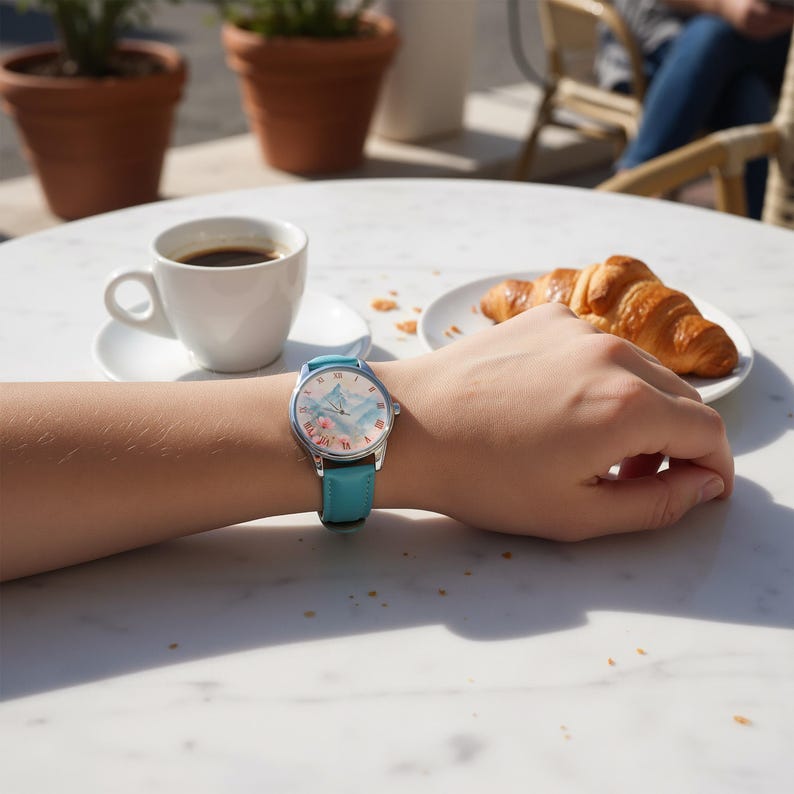 K&ouml;nnte beinhalten: Silberne Armbanduhr mit hellblauem Lederarmband. Das Zifferblatt zeigt eine Berglandschaft mit rosa floralen Akzenten. Die Uhr befindet sich an einem Handgelenk, mit Kaffee und einem Croissant im Hintergrund.