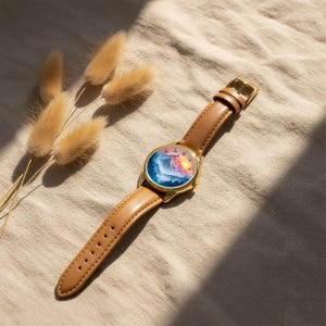 May include: A gold-tone wristwatch with a brown leather band. The watch face features a colorful mountain landscape design. The watch is lying on a beige surface, with dried ornamental grass in the background.