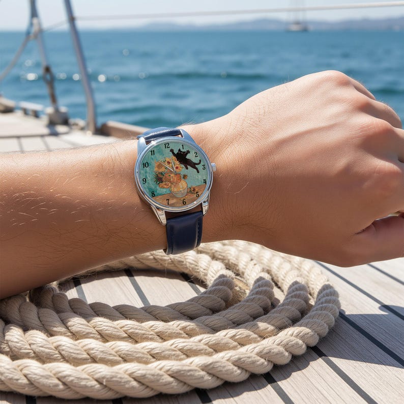 Puede incluir: Un reloj de pulsera con una correa de cuero azul y una caja plateada. La esfera del reloj presenta una pintura de un gato negro y flores naranjas. El reloj est&aacute; en una mu&ntilde;eca, con una cuerda n&aacute;utica en primer plano.