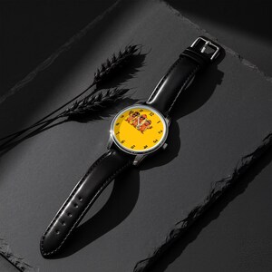 May include: A wristwatch with a black leather strap and a round silver-tone case. The watch face is bright yellow with black numerals and features a central graphic illustration of three anthropomorphic hot dogs wearing sunglasses.