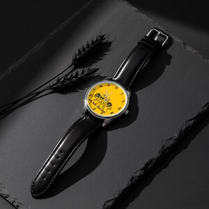 Puede incluir: Un reloj de pulsera con correa de cuero negro y caja redonda de tono plateado. La esfera es de color amarillo brillante con n&uacute;meros negros y presenta una ilustraci&oacute;n gr&aacute;fica de tres personajes de dibujos animados con gafas de sol.