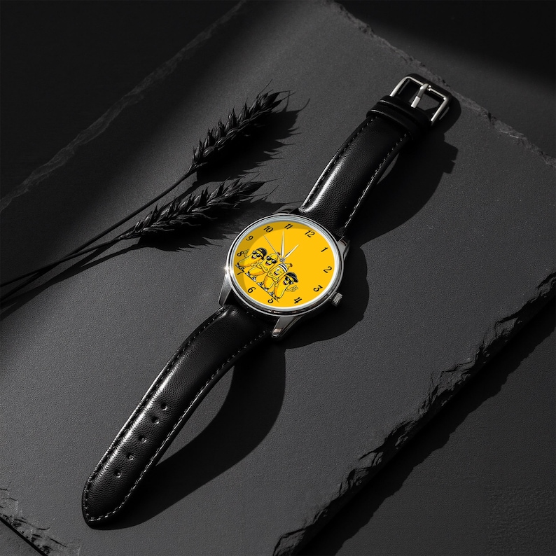 Puede incluir: Un reloj de pulsera con correa de cuero negro y caja redonda de tono plateado. La esfera es de color amarillo brillante con n&uacute;meros negros y presenta una ilustraci&oacute;n gr&aacute;fica de tres personajes de dibujos animados con gafas de sol.