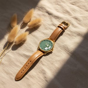 May include: A gold-tone wristwatch with a green face featuring a cartoon illustration. The watch has a brown leather strap and a gold buckle. The watch is set against a neutral background with dried flowers.