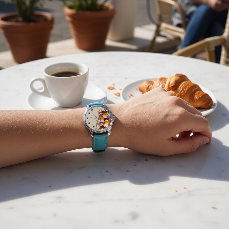 May include: A round wristwatch with a light blue leather band. The watch face features a collection of cartoon cats in various colors. The watch is on a person's wrist, with a cup of coffee and a croissant in the background.