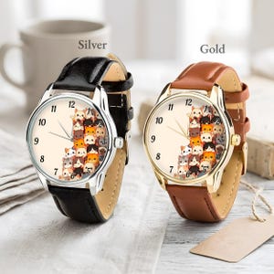 May include: Two wristwatches with cream-colored faces and black leather bands. One watch has a silver case, the other a gold case. The watch faces feature a pattern of stacked cartoon cats. The word "Silver" is above the silver watch, and "Gold" is above the gold watch.