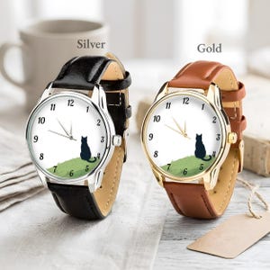 Op de afbeelding: Twee polshorloges, &eacute;&eacute;n zilver en &eacute;&eacute;n goud. Elk horloge heeft een witte wijzerplaat met zwarte cijfers en een zwart katten silhouet op een groene heuvel. Het zilveren horloge heeft een zwarte leren band, en het gouden horloge heeft een bruine leren band.