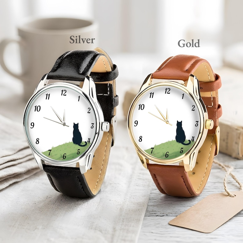 Op de afbeelding: Twee polshorloges, &eacute;&eacute;n zilver en &eacute;&eacute;n goud. Elk horloge heeft een witte wijzerplaat met zwarte cijfers en een zwart katten silhouet op een groene heuvel. Het zilveren horloge heeft een zwarte leren band, en het gouden horloge heeft een bruine leren band.