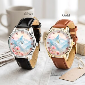 K&ouml;nnte beinhalten: Zwei Armbanduhren. Eine hat ein silbernes Geh&auml;use und ein schwarzes Armband, die andere ein goldenes Geh&auml;use und ein braunes Armband. Beide haben ein Berg- und Blumendesign auf dem Zifferblatt mit r&ouml;mischen Ziffern. Die W&ouml;rter "Silver" und "Gold" sind sichtbar.